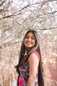 A vertical portrait of a young woman posing for the camera with white blossoms behind her.