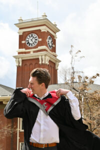 A vertical photo of a young man in front of a clock tower looking off to the side as wind blows in his hair.