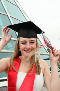 A vertical portrait of a young woman with a black grad cap on with her hair blowing in the wind.