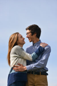 A vertical photo of a young couple in each others arms smiling at each other with the blue sky behind them.