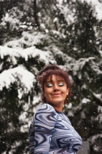 A vertical photo of a young woman smiling at the camera with her eyes closed and snow falling around her.